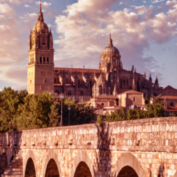Fotografia de Salamanca: grande ponte de pedras com design romano/grego, abaixo dela um riacho e ao fundo uma bela igreja.